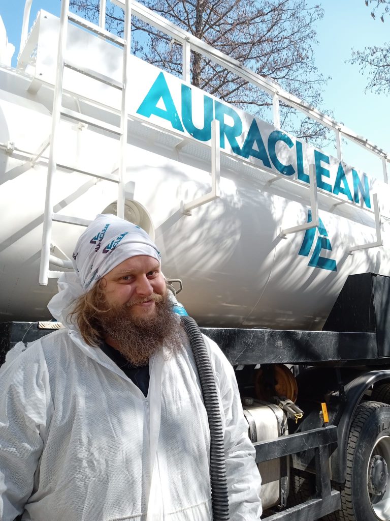 Työntekijä Valtteri Urponen seisoo AuraCleanin imuauton vieressä valkoisessa haalarissa firman logolla varustettu huivi päässään ja hymyilee.