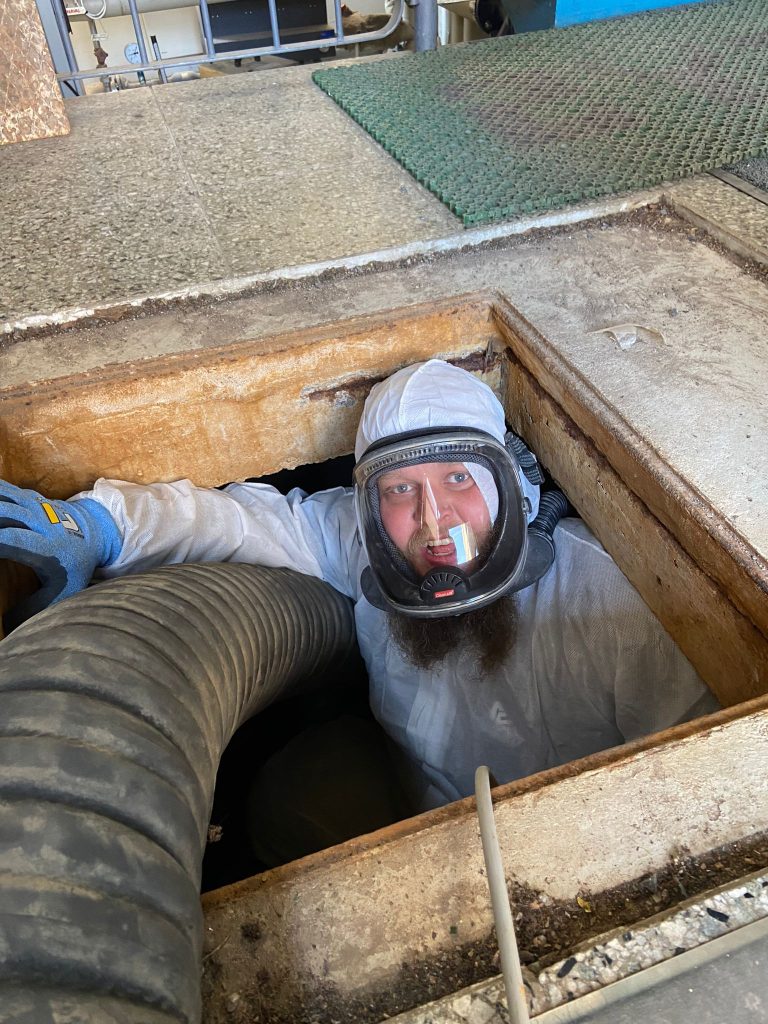 Työntekijä Valtteri Urponen on betonikuopassa ison harmaan letkun kanssa pukeutuneena suojapukuun ja naamariin.